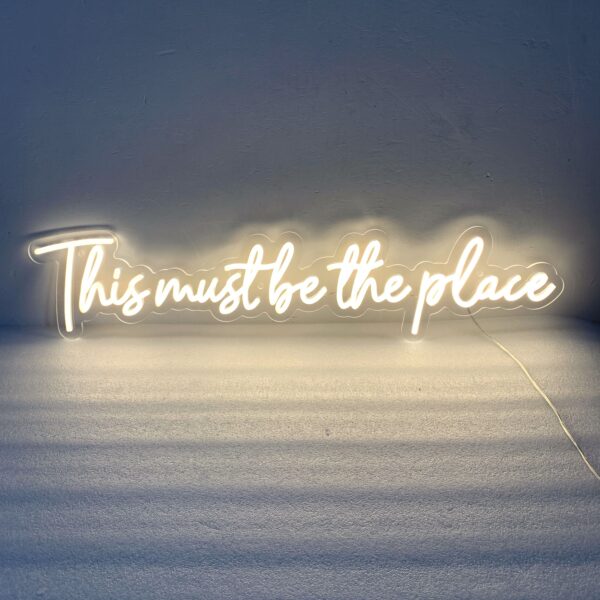 A glowing neon sign with the phrase 'This Must Be The Place' in vibrant colors, evoking warmth and comfort.